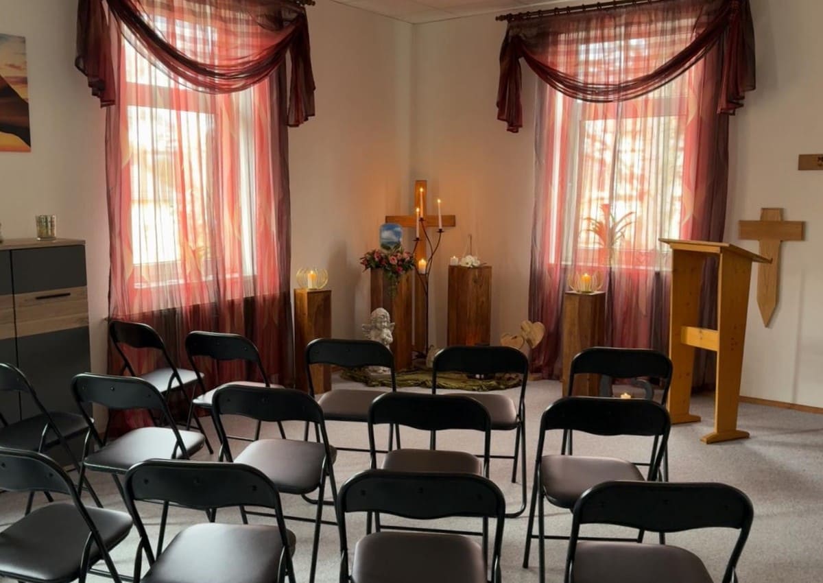 Innenansicht des Abschiedsraums im Bestattungshaus Sternenblick: Stuhlreihen führen zu einer warm beleuchteten Gedenkstätte mit Urne, Blumen und Kerzen, umrahmt von Fenstern mit rot-cremefarbenen Gardinen.